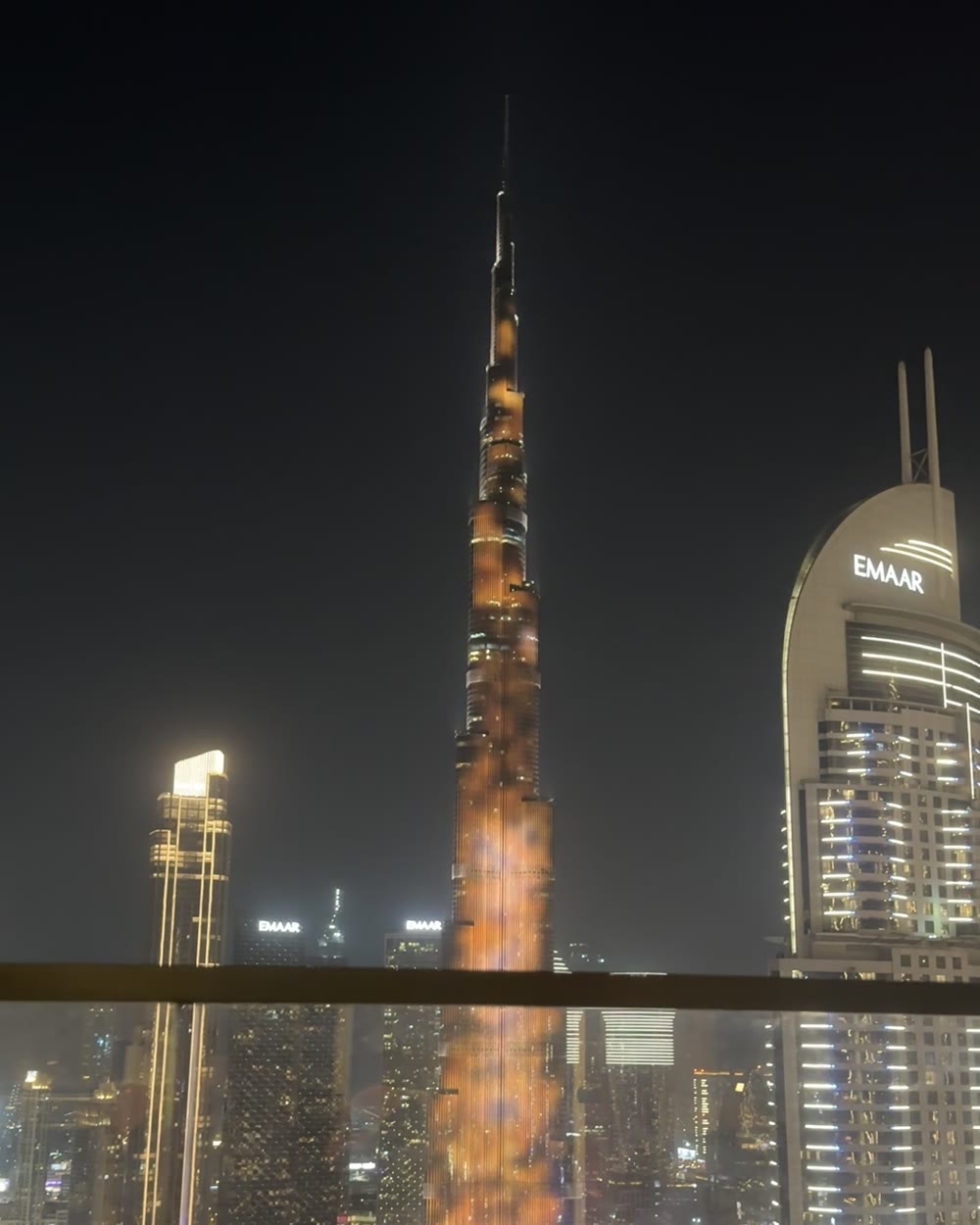 The Burj Khalifa lit up orange at night, seen over a balcony rail with neighboring towers